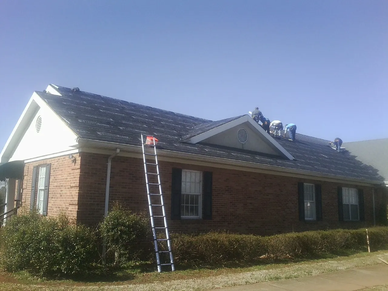 Crew tearing shingles off a brick ranch roof with a ladder set — Metal Roof Repair work by our crew in Kidder