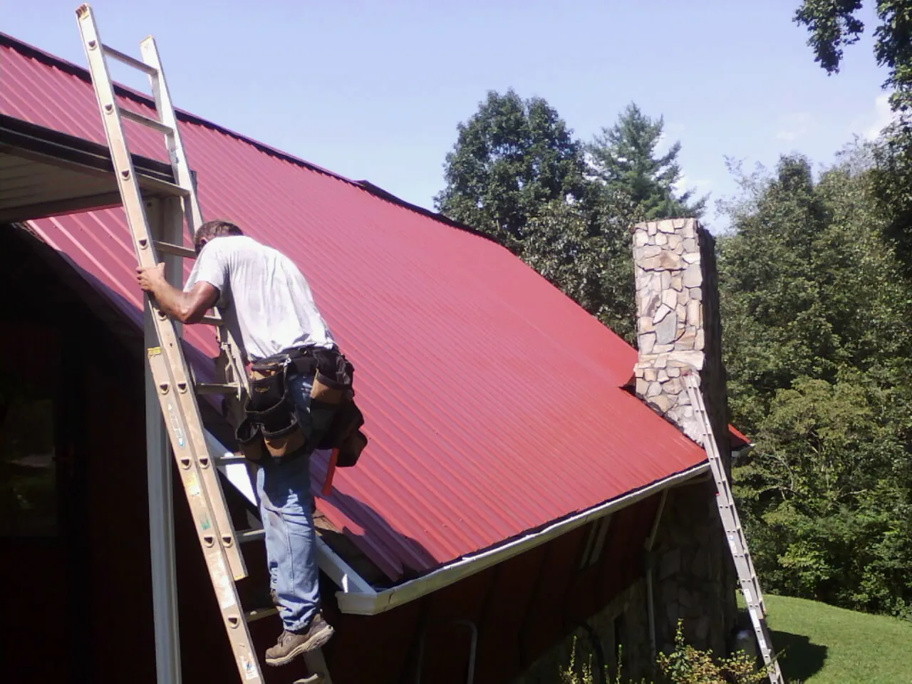 Roofer on a ladder beside a red metal roof and stone chimney — Soffit & Fascia Repair by Roofing Brothers in Kidder